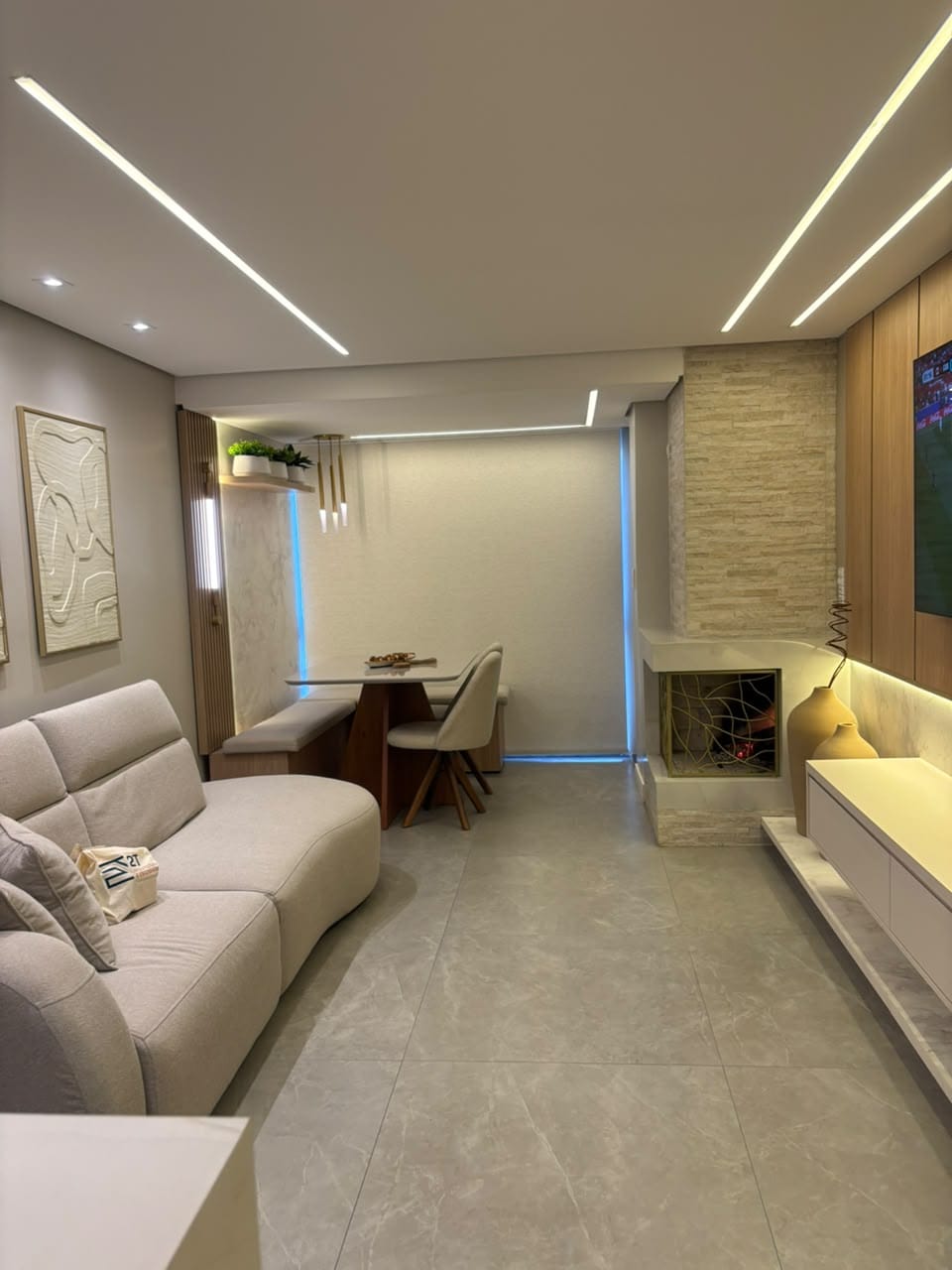 Sala de estar com sofá claro, painel de TV amadeirado e jantar ao fundo