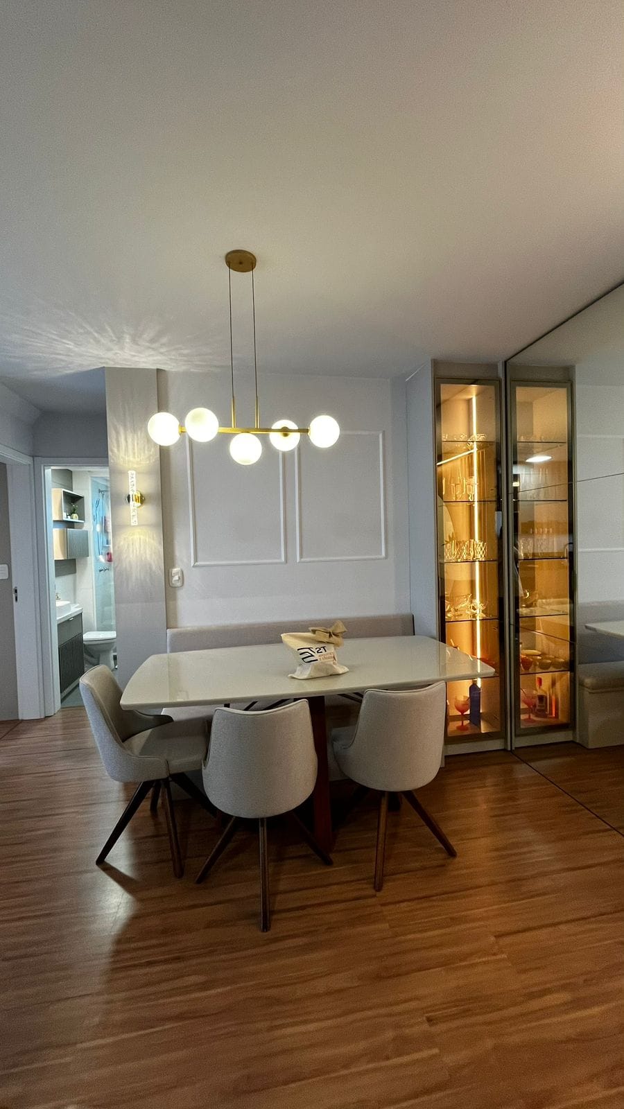 Sala de jantar com mesa clara, pendente de globos e cristaleira iluminada