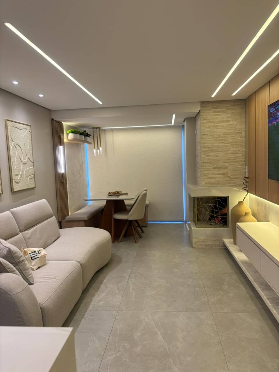 Sala de estar com sofá claro, painel de TV amadeirado e jantar ao fundo